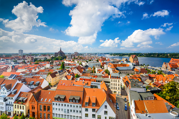 Blick von oben auf die roten Ziegeldächer der Rostocker Innenstadt mit der Marienkirche im Hintergrund und der Warnow am rechten Bildrand.