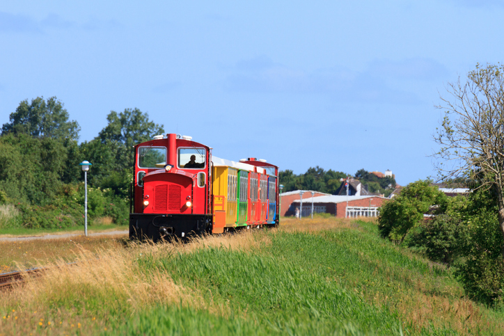 Eine rote Diesellok mit bunten Passagierwaggons fährt auf einer Schiene durch die grüne Wiesen- und Dünenlandschaft der Nordseeinsel Langeoog.