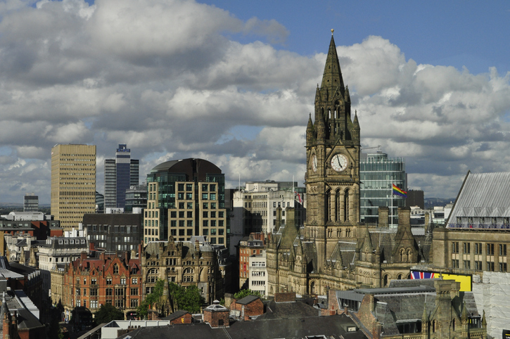 Blick über die Dächer von Manchester auf den hohen, gotischen Turm des Rathauses (Town Hall) vor einer Kulisse aus modernen Bürogebäuden.