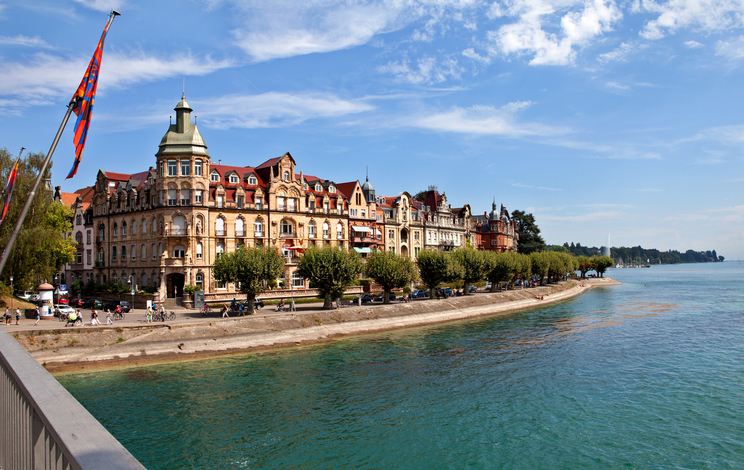Historische, herrschaftliche Gebäude mit roten Dächern erstrecken sich entlang des glitzernden Seeufers in Konstanz.