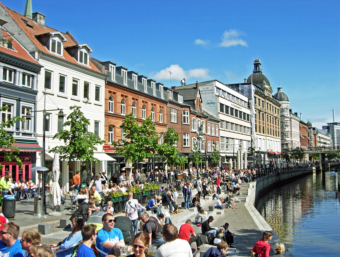 Viele Menschen sitzen in Cafés und auf Treppen am Kanal Aarhus Å unter blauem Himmel in der Innenstadt.