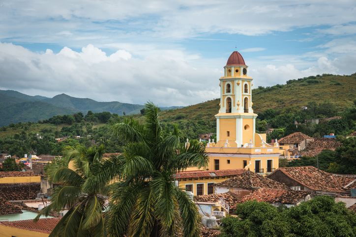 Ein gelber Glockenturm einer Kirche überragt die roten Ziegeldächer und Palmen in der historischen Stadt Trinidad auf Kuba.