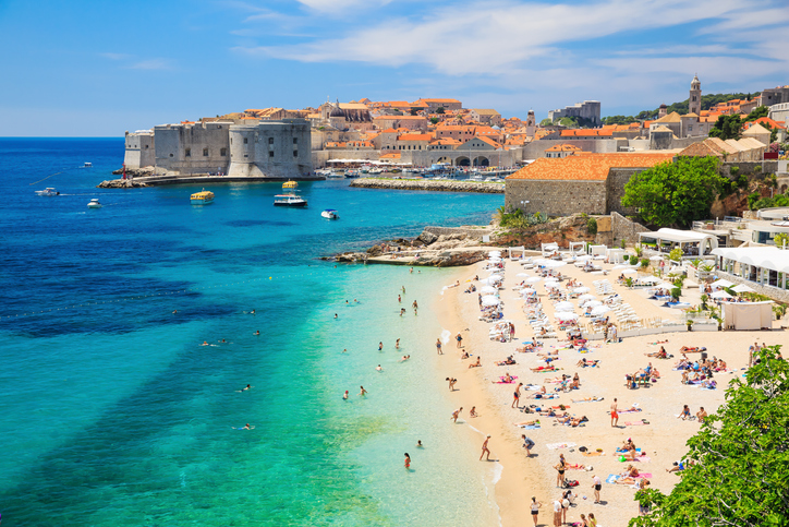 Ein belebter Sandstrand mit weißen Sonnenschirmen vor der berühmten mittelalterlichen Stadtmauer und den roten Dächern von Dubrovnik.