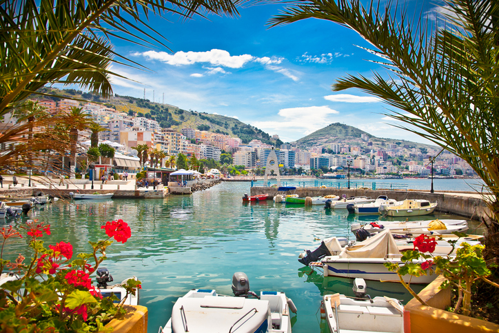 Kleine Boote liegen im türkisfarbenen Wasser eines Hafens in Saranda, umrahmt von Palmen und der albanischen Küstenstadt.