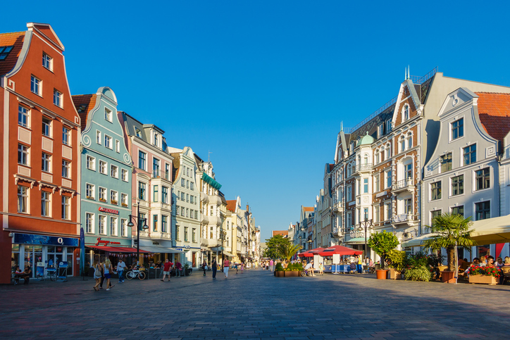 Eine weite Fußgängerzone mit historischen Häuserfronten, Cafés und Menschen, die unter einem wolkenlosen blauen Himmel spazieren.