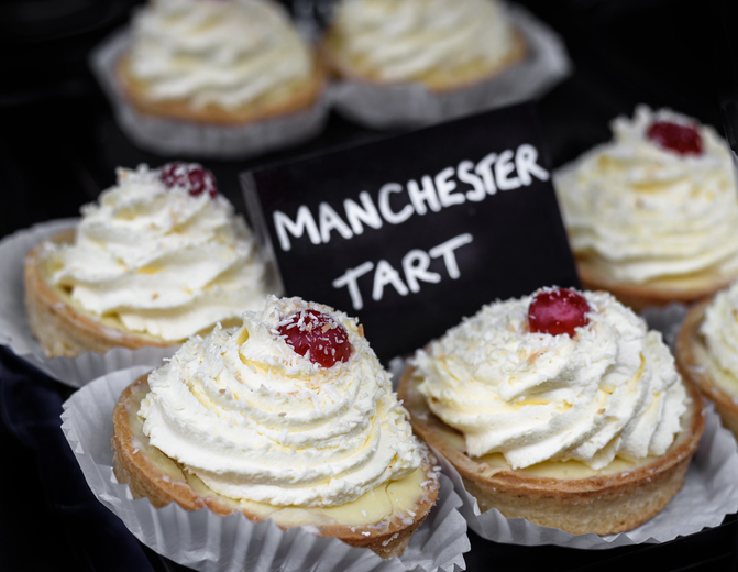 Mehrere kleine Törtchen mit einer Haube aus weißer Creme, Kokosraspeln und einer roten Kirsche obendrauf, mit einem Preisschild "Manchester Tart".