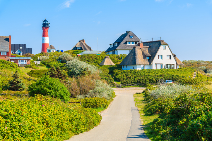 Ein rot-weißer Leuchtturm ragt hinter einer Dünenlandschaft mit grünen Büschen und charakteristischen Häusern mit Reetdach hervor.