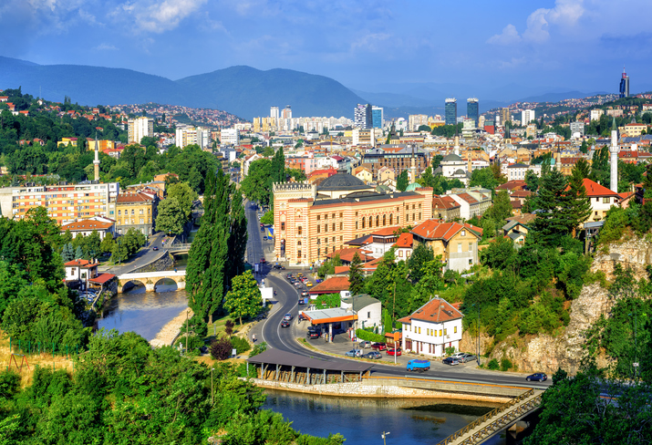 Blick über den Fluss Miljacka auf die vielfältige Architektur Sarajevos mit der Nationalbibliothek und zahlreichen Minaretten.