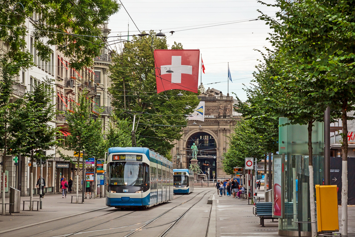 Ein blaues Zürcher Tram fährt durch die belebte Bahnhofstrasse unter einer wehenden Schweizer Flagge.
