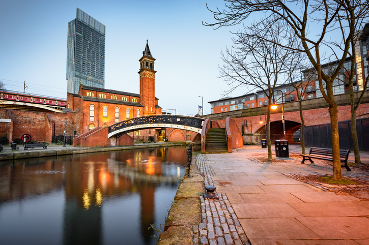 Ein beleuchteter Backsteinbau mit Turm an einem ruhigen Kanal, davor eine kleine schwarze Bogenbrücke und eine Uferpromenade.