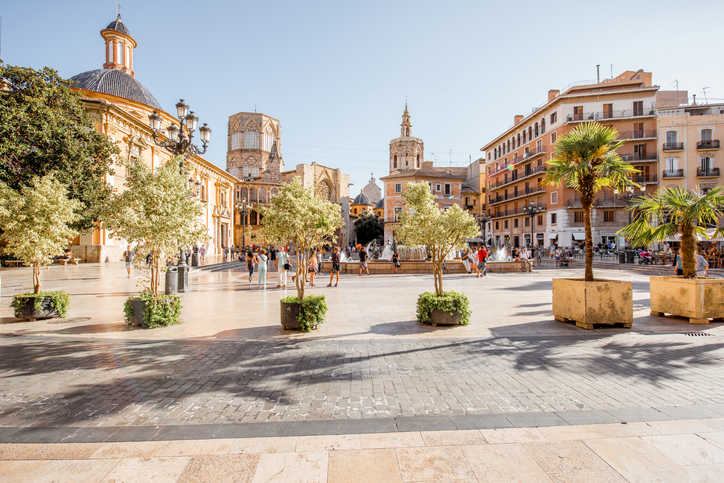 Ein sonniger Platz in Valencia mit Bäumen, Passanten und historischen Kirchengebäuden im Hintergrund.