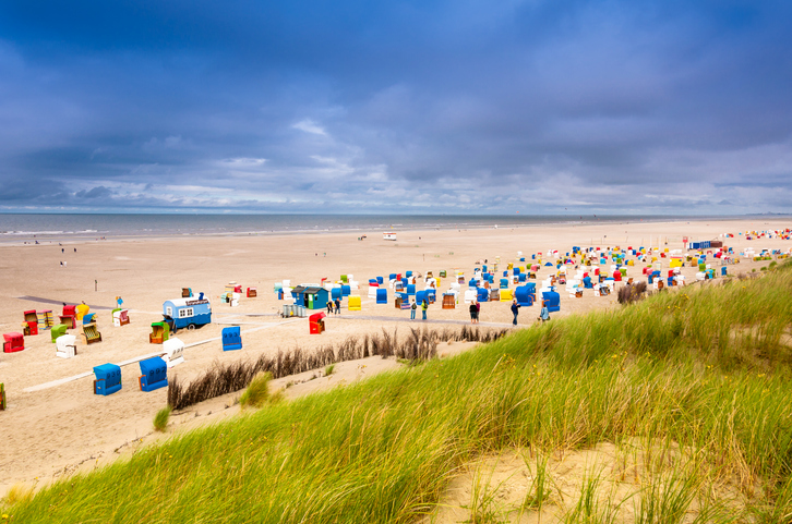 Ein weiter Sandstrand auf der Nordseeinsel Juist mit zahlreichen bunten Strandkörben, grünen Dünen im Vordergrund und einem bewölkten Himmel.
