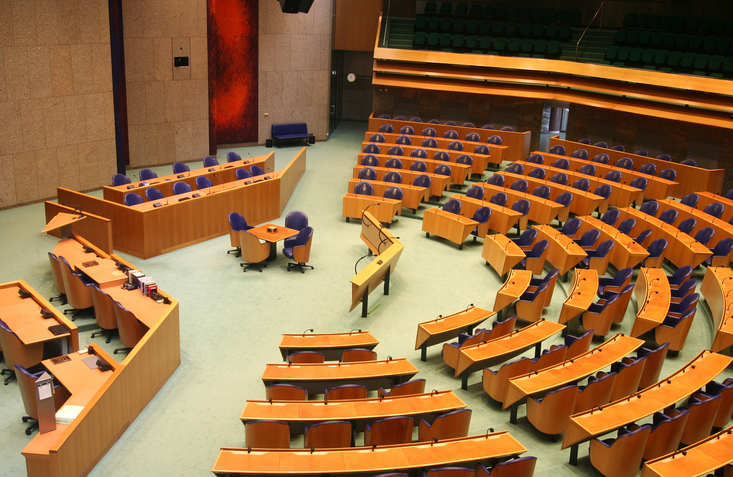 Blick von oben in den halbkreisförmigen Sitzungssaal des niederländischen Parlaments mit blauen Stühlen und hellen Holztischen.