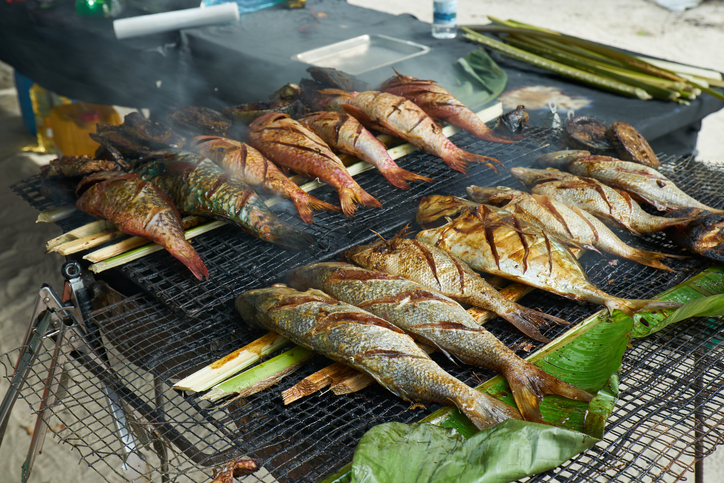 Mehrere ganze Fische werden auf einem Grill über offenem Feuer an einem Sandstrand zubereitet.
