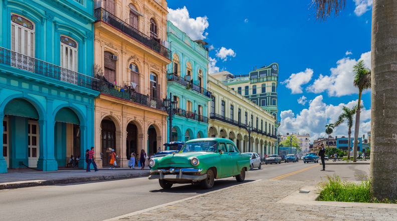 Ein grüner Oldtimer fährt durch eine Straße in Havanna, gesäumt von farbenfrohen Häuserfassaden im Kolonialstil.