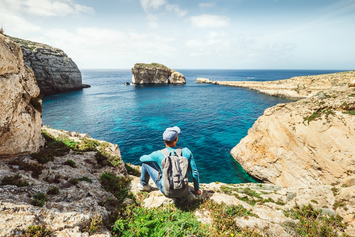Ein Wanderer mit Rucksack sitzt auf einer Klippe in Gozo und blickt auf den markanten Fungus Rock im Meer.