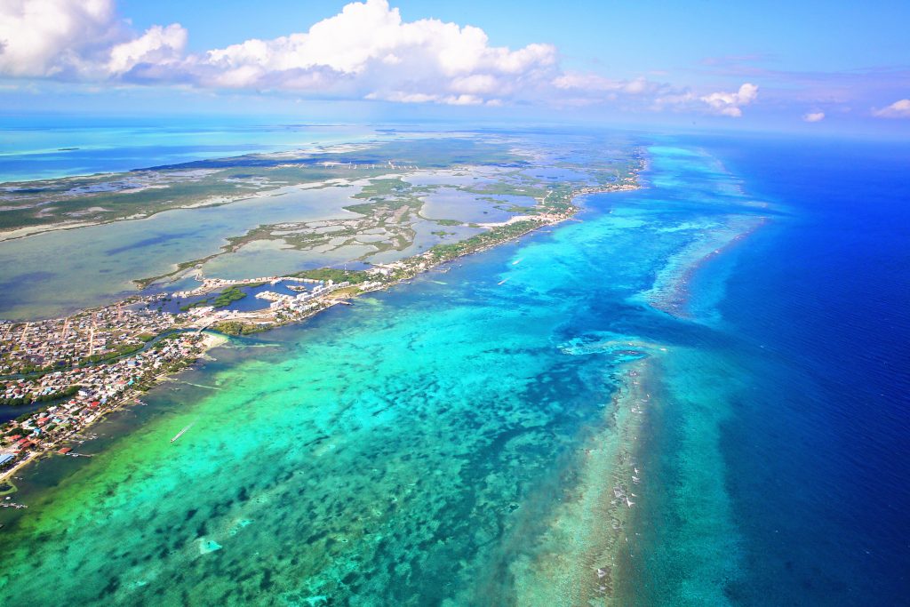 Ein Luftbild zeigt eine langgezogene tropische Insel mit flachen Lagunen, die von tiefblauem Meer umgeben ist.