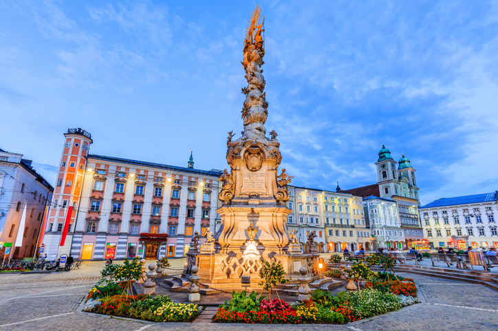 Die kunstvolle Dreifaltigkeitssäule steht beleuchtet auf einem weiten Platz, umgeben von historischen Fassaden bei blauem Abendhimmel.