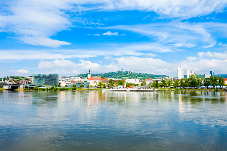Ein weiter Blick auf den Flusslauf zeigt die Stadtkulisse, ein Ausflugsschiff und die grüne Hügellandschaft unter einem heiteren Sommerhimmel.