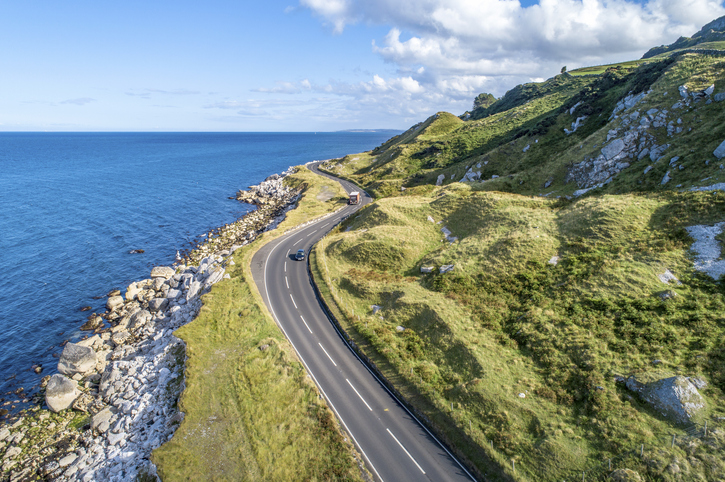 Eine sich windende Straße führt entlang der grünen Hügel und der felsigen Küstenlinie Nordirlands direkt am Meer entlang.