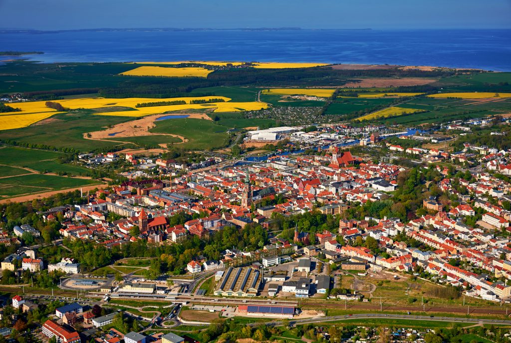 Eine weite Luftaufnahme zeigt das gesamte Stadtgebiet von Greifswald, umgeben von grünen Feldern und Rapsblüten.