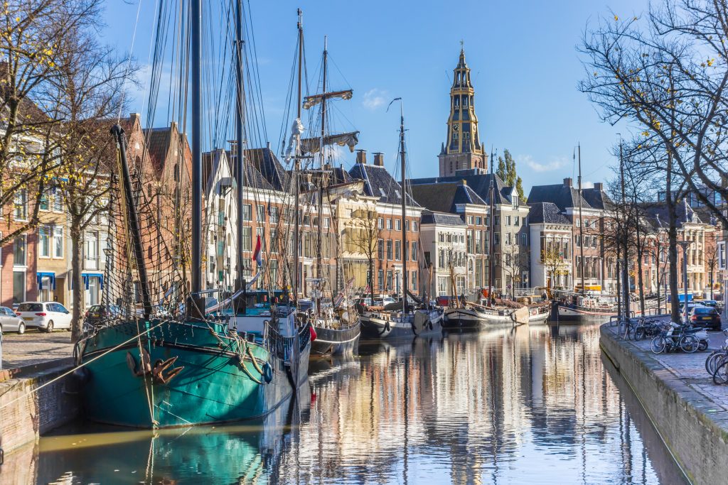 Im Kanal vor einer historischen Häuserreihe liegen alte Segelschiffe vor Anker, im Hintergrund ragt der Martiniturm empor.