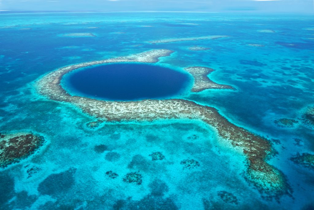 Eine Luftaufnahme zeigt das tiefblaue, kreisförmige Great Blue Hole inmitten eines flachen, türkisfarbenen Korallenriffs.