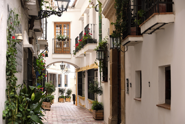 Eine enge, weiß getünchte Gasse in Marbella, geschmückt mit grünen Pflanzen, Blumen in Kästen an den Balkonen und historischen Laternen.