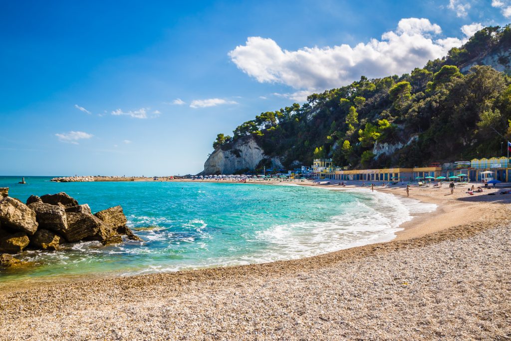 Ein Kiesstrand mit türkisfarbenem Wasser, umgeben von bewaldeten Klippen unter einem strahlend blauen Himmel.