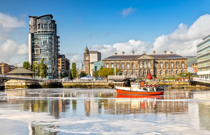 Ein modernes Hochhaus und ein historisches Gebäude stehen sich am Ufer des Lagan in Belfast gegenüber, während ein rotes Boot im Wasser liegt.