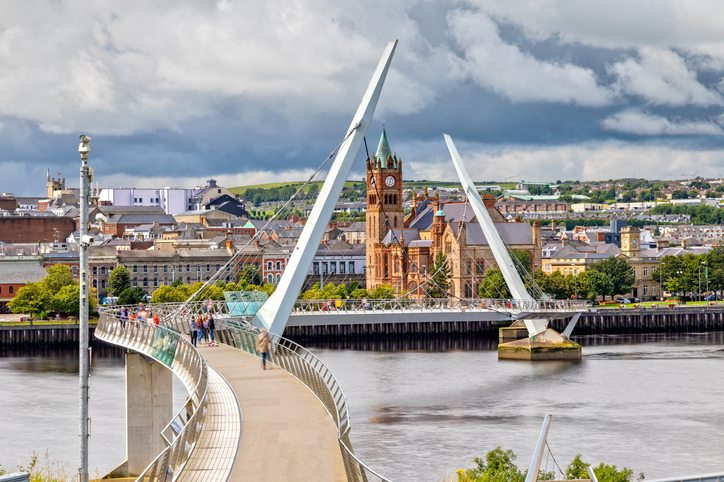 Die geschwungene, moderne Fußgängerbrücke „Peace Bridge“ führt über den Fluss Foyle, im Hintergrund ist das historische Rathaus zu sehen.
