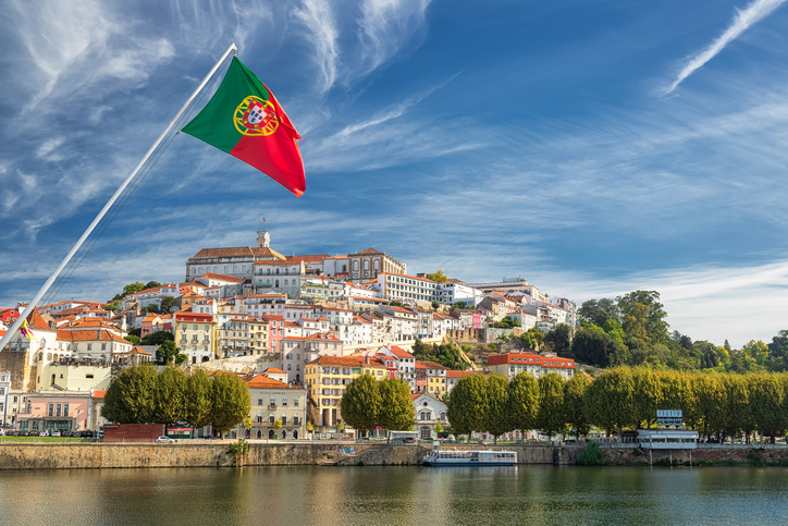 Die portugiesische Nationalflagge weht im Wind vor der historischen Kulisse der Stadt Coimbra am Flussufer.