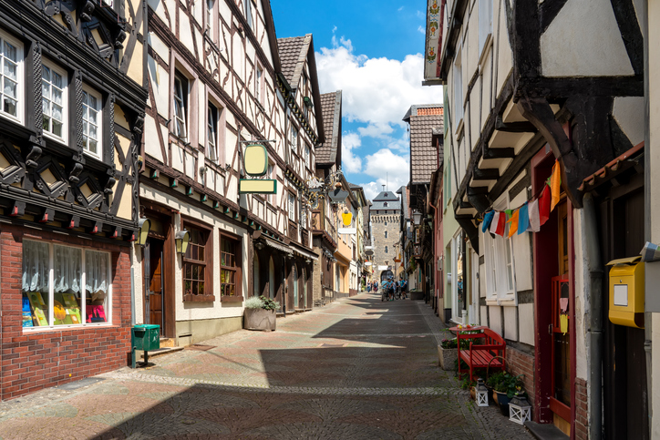 Eine schmale, gepflasterte Gasse gesäumt von traditionellen Fachwerkhäusern führt auf einen alten Turm im Hintergrund zu.