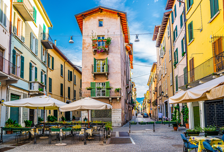 Eine enge, charmante Straße in Brescia mit farbenfrohen Gebäuden, die von Sonnenschirmen und Außenbestuhlung gesäumt wird.