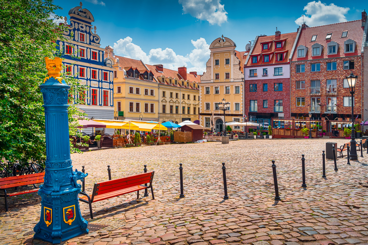 Ein leerer, gepflasterter Platz mit farbenfrohen Giebelhäusern und einer auffälligen blauen historischen Wasserpumpe.