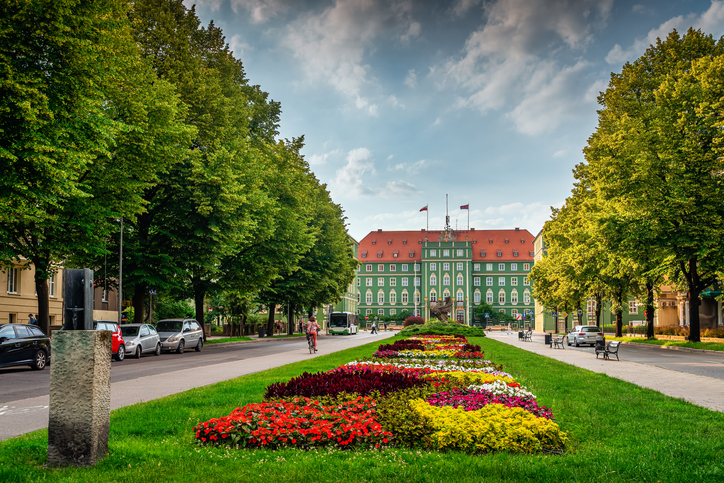 Eine von Bäumen gesäumte Straße führt auf ein imposantes grünes Gebäude zu, davor ein bunt bepflanztes Blumenbeet.
