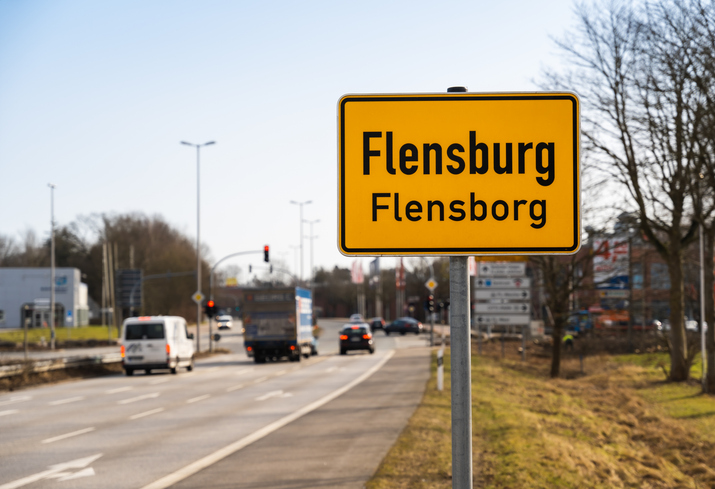 Ein gelbes Ortsschild mit der Aufschrift „Flensburg“ und der dänischen Bezeichnung „Flensborg“ steht am Rand einer Straße mit beginnendem Verkehr.