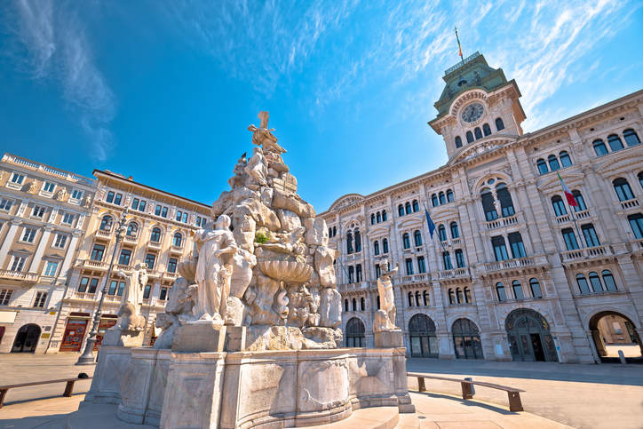 Ein kunstvoller, weißer Marmorbrunnen vor der prächtigen historischen Fassade des Hauptgebäudes auf der Piazza Unità d'Italia.