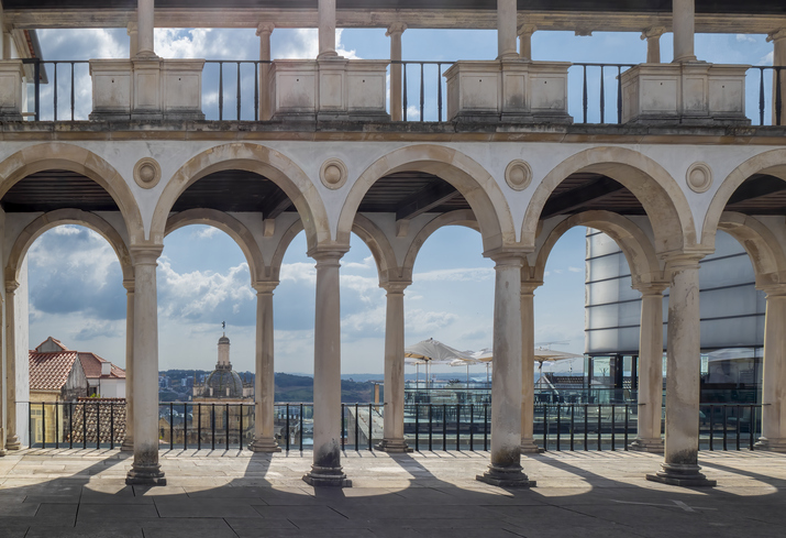 Blick durch die klassischen steinernen Bögen der Universität von Coimbra auf den Stadtbereich und die Hügellandschaft.