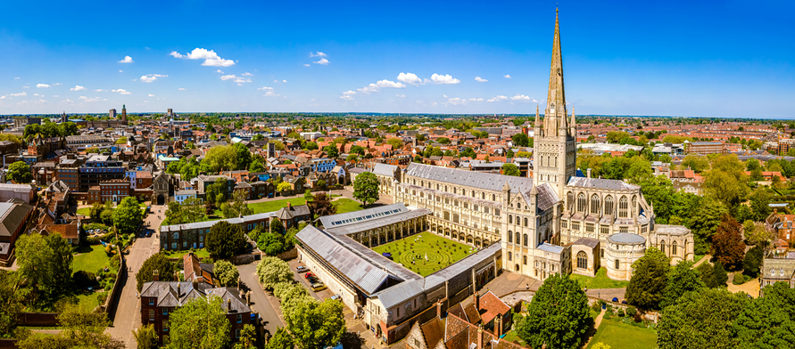 Ein heller Blick auf die Kathedrale von Norwich mit ihren weitläufigen Gärten und dem charakteristischen Turm unter blauem Himmel.
