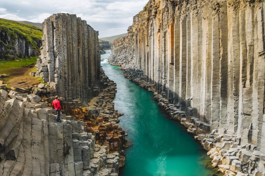 Ein Wanderer in roter Jacke steht auf einer Klippe über einem Fluss, der durch eine tiefe Schlucht aus hohen Basaltsäulen fließt.