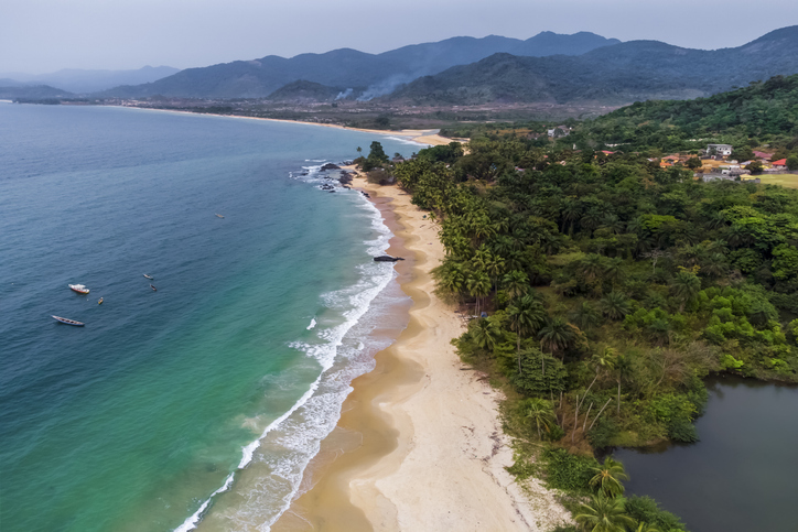 Ein langer, goldener Sandstrand in Sierra Leone säumt das türkisfarbene Meer, während dichte grüne Bäume an die Küste grenzen.