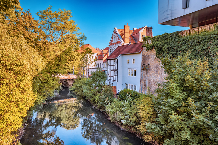 Ein ruhiger Fluss fließt durch eine grüne, baumreiche Landschaft mit historischen Gebäuden im Hintergrund.
