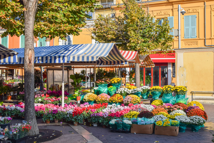 Farbenfrohe Blumenstände stehen vor gelben Häuserfassaden auf einem Markt in Nizza.
