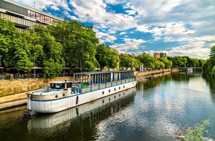 Ein flaches Fahrgastschiff fährt auf dem Neckar durch die grüne Uferlandschaft von Heilbronn unter einem wolkenverhangenen Himmel.