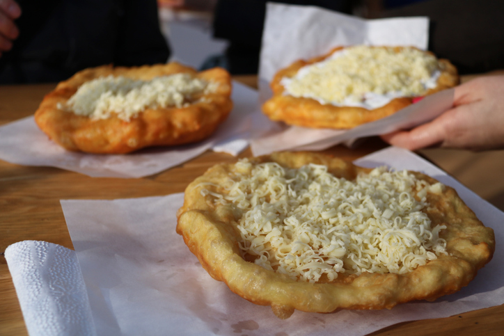 Drei frisch gebackene Lángos mit geriebenem Käse liegen auf Papier auf einem Holztisch.