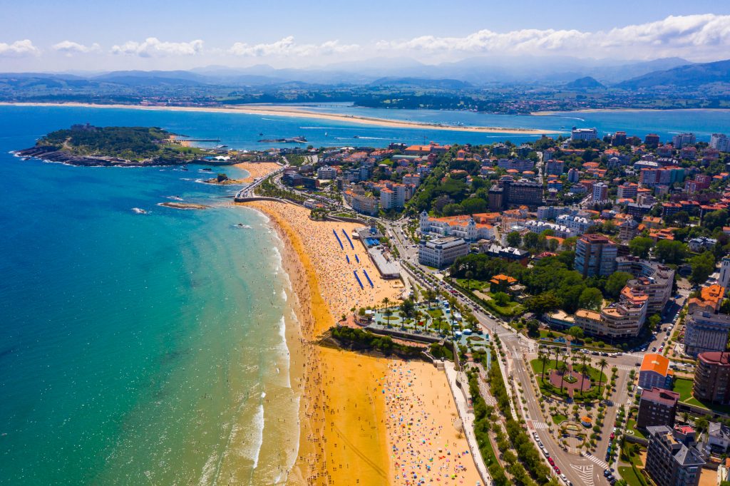 Eine weite Luftaufnahme zeigt einen langen, goldenen Sandstrand, der von zahlreichen Menschen besucht wird, mit der Stadt im Hintergrund.