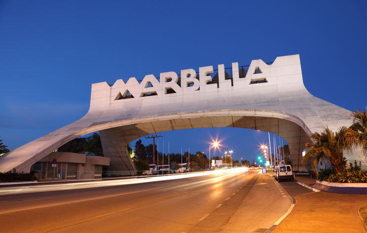 Das ikonische, moderne architektonische Eingangstor mit dem Schriftzug „MARBELLA“ bei Einbruch der Nacht, mit Lichtspuren vorbeifahrender Autos.
