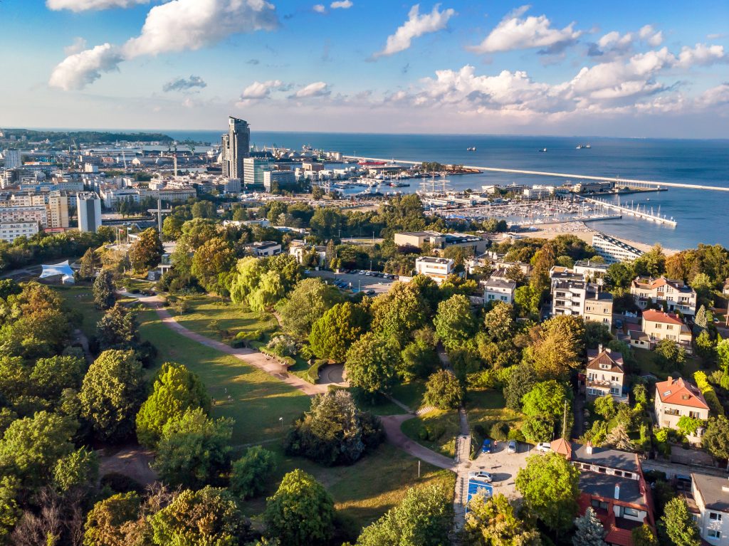 Ein grüner Park mit Bäumen im Vordergrund bietet einen weitläufigen Ausblick über Gdynia, den Hafen und die Ostsee.