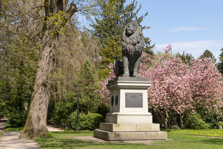 Eine bronzene Löwenstatue thront auf einem hohen Steinsockel inmitten einer grünen Parkanlage mit blühenden rosa Bäumen im Hintergrund.